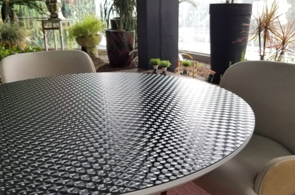 Uma mesa redonda com tampo preto texturizado em padrão de losangos fica entre duas cadeiras de cor clara em uma sala iluminada.