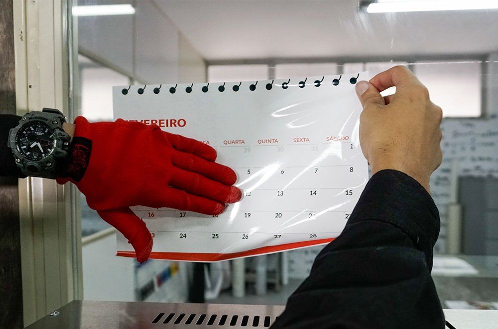 Uma pessoa com uma luva vermelha segura um calendário de fevereiro com encadernação em espiral contra uma superfície limpa em uma área de trabalho.