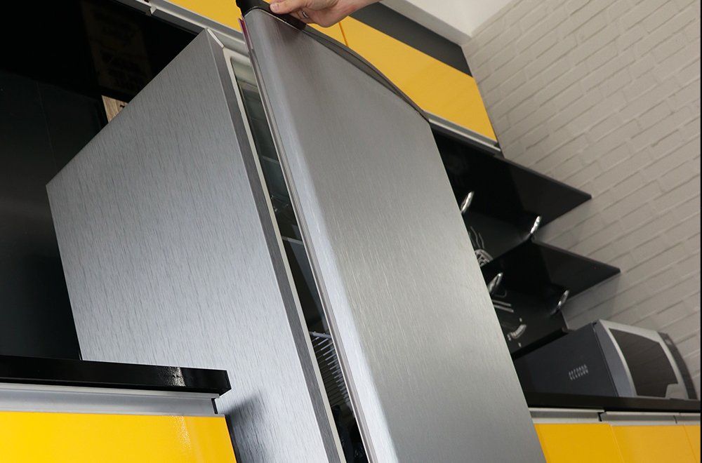 Visão aproximada de uma mão abrindo a porta prateada de uma geladeira em uma cozinha com armários amarelos e pretos.