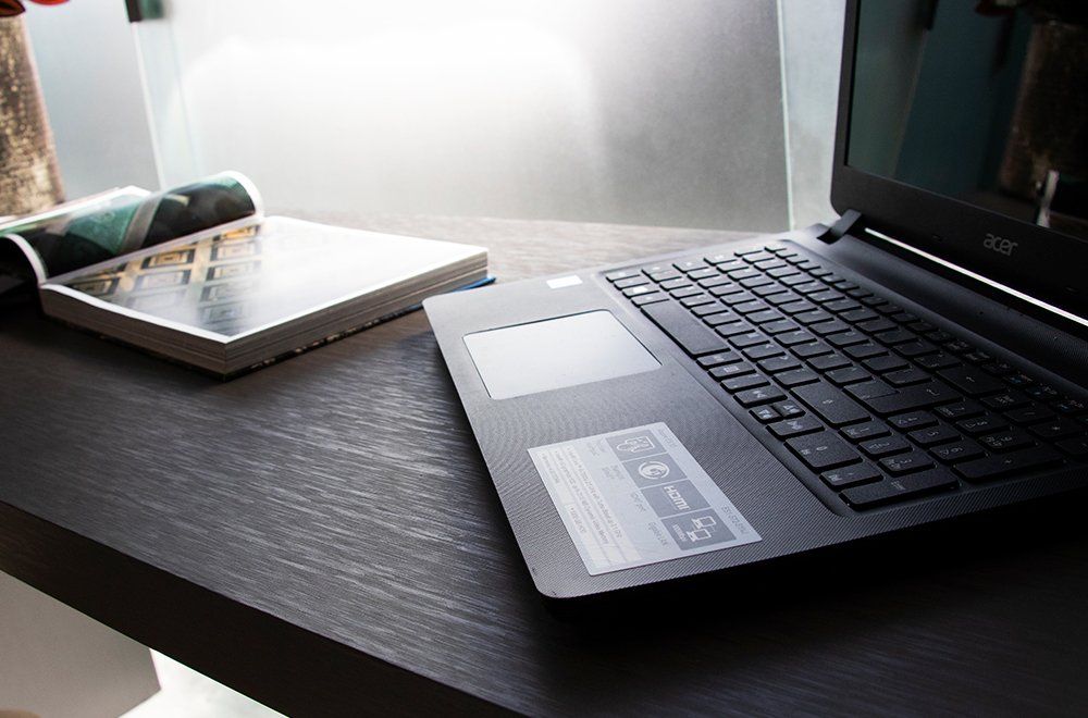 Um laptop preto está sobre uma mesa de madeira escura ao lado de um livro aberto, iluminado por uma luz natural intensa.