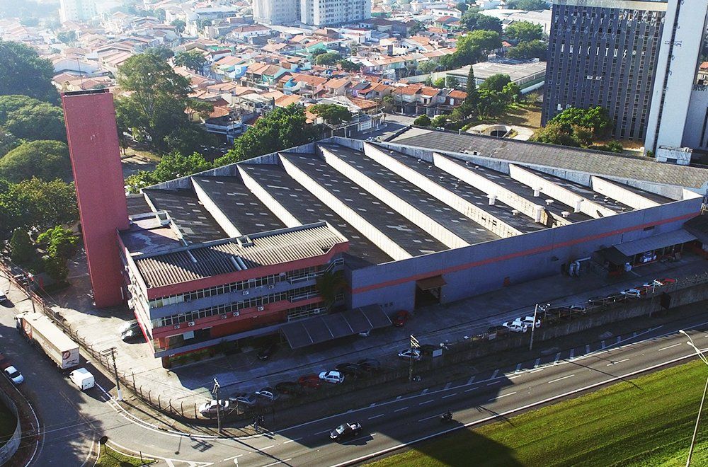 Vista aérea de uma fábrica com uma alta chaminé vermelha e um telhado em forma de dente de serra, localizada ao lado de uma estrada e edifícios residenciais.