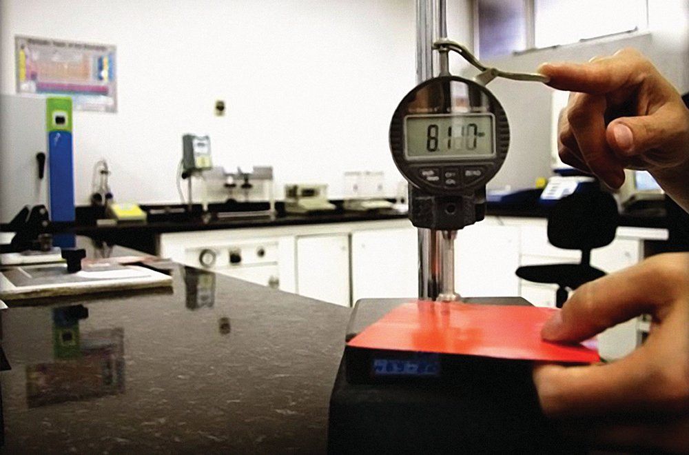 Uma pessoa utiliza um medidor de profundidade digital para medir a espessura de um material vermelho em formato quadrado em um laboratório.
