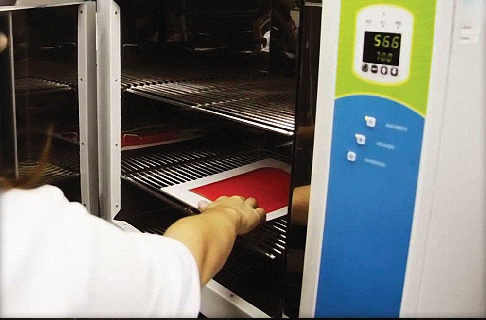 Uma pessoa coloca uma serigrafia retangular vermelha em uma prateleira dentro de uma incubadora de laboratório com um painel de controle digital.