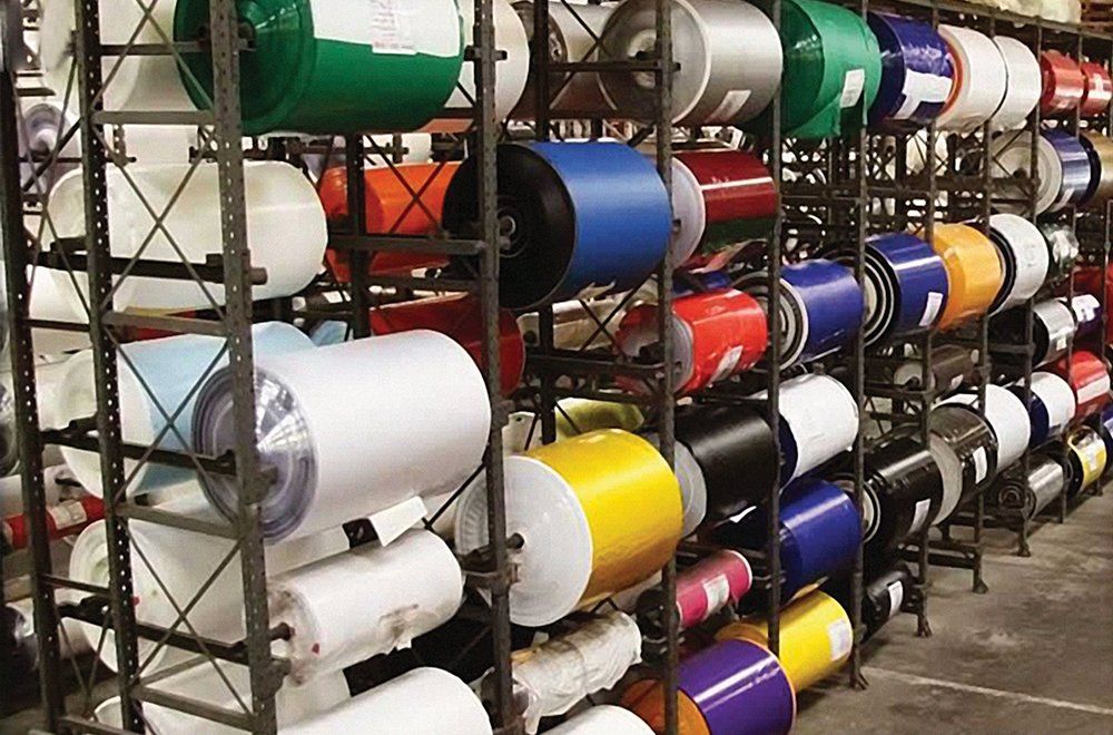 Sistema de estantes metálicas industriais repleto de inúmeros rolos coloridos de material plástico ou vinílico em um ambiente de armazém.