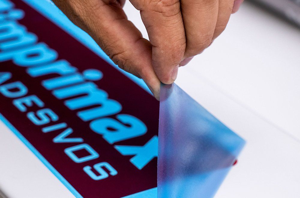 Uma mão retirando uma película protetora transparente de uma superfície branca com o logotipo azul da Primax Adesivos impresso nela.