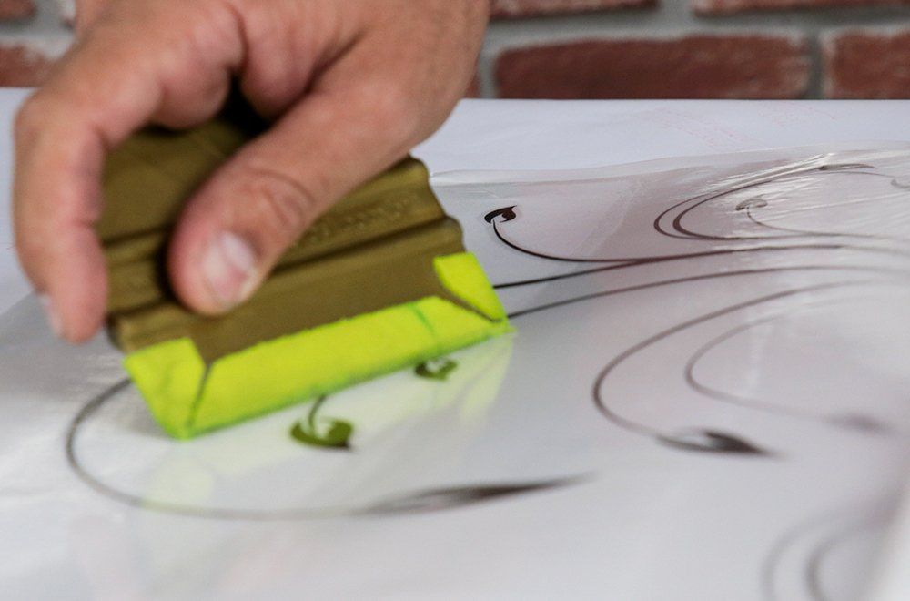 Uma mão utiliza uma espátula verde e amarela para alisar um decalque decorativo de vinil sobre uma superfície branca plana.