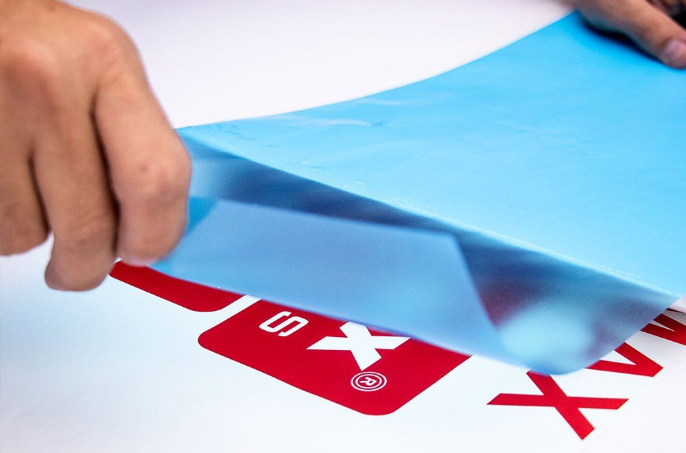 Mãos retiram uma película protetora azul translúcida de uma superfície branca com texto e logotipos impressos em vermelho.