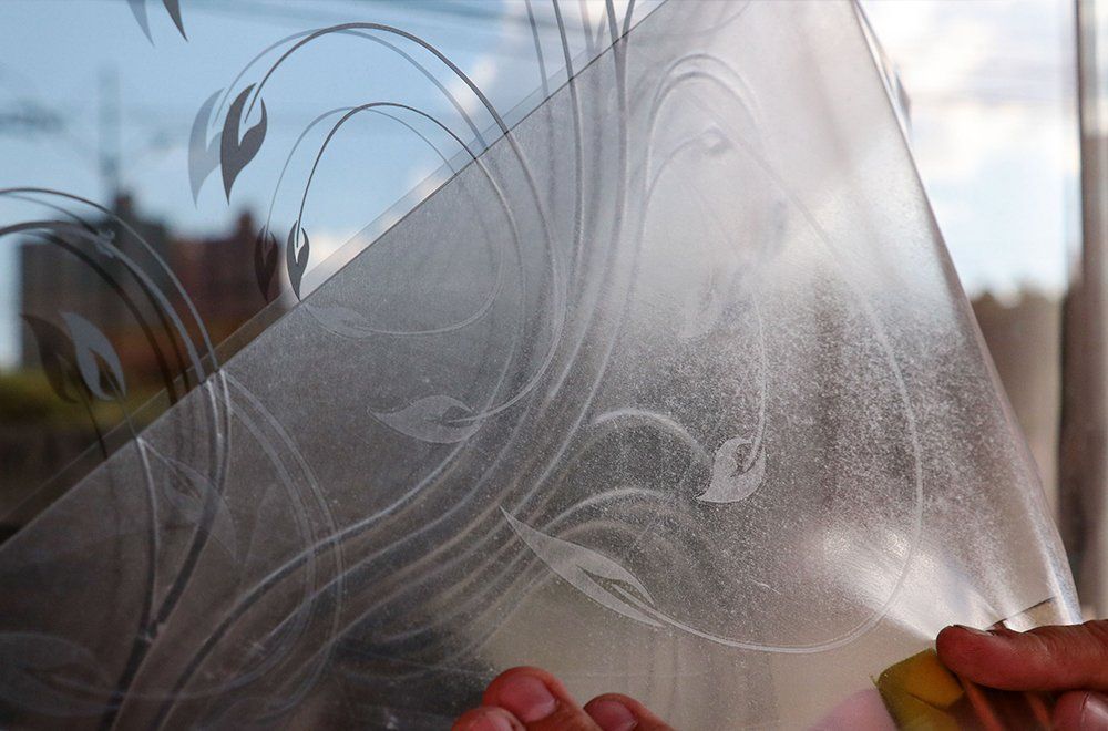 Mãos segurando um pedaço de película decorativa fosca para janela com um padrão floral branco em espiral.