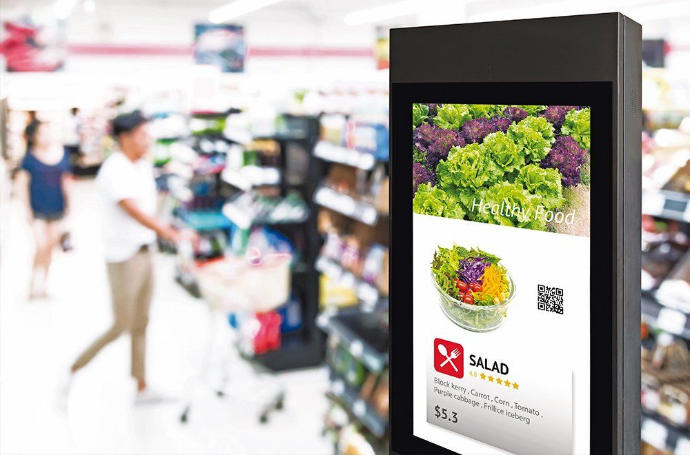 Um quiosque digital exibe um anúncio de salada em um corredor desfocado de um supermercado, onde pessoas estão fazendo compras.