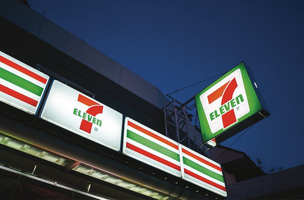 A placa externa e o logotipo de uma loja 7-Eleven iluminados contra um céu azul escuro ao entardecer.