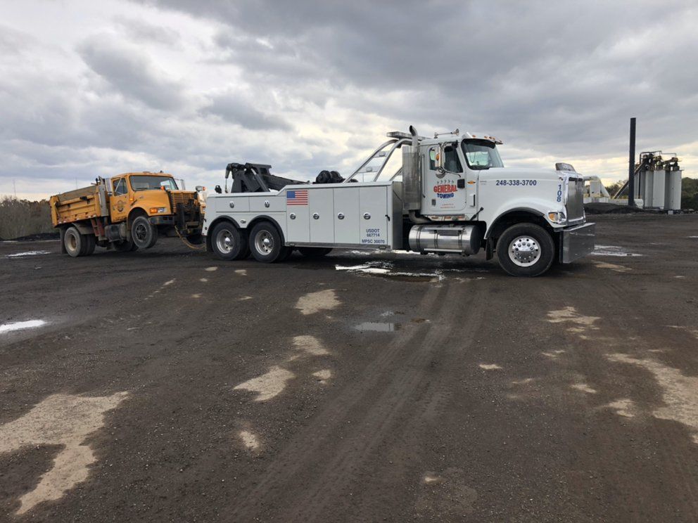 White tow truck towing a yellow dump truck on a dark, wet surface under a cloudy sky.