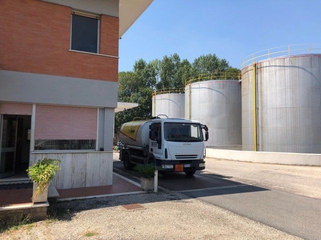In una giornata di sole, un'autocisterna bianca è parcheggiata accanto a un edificio in mattoni e a grandi serbatoi di stoccaggio in metallo.