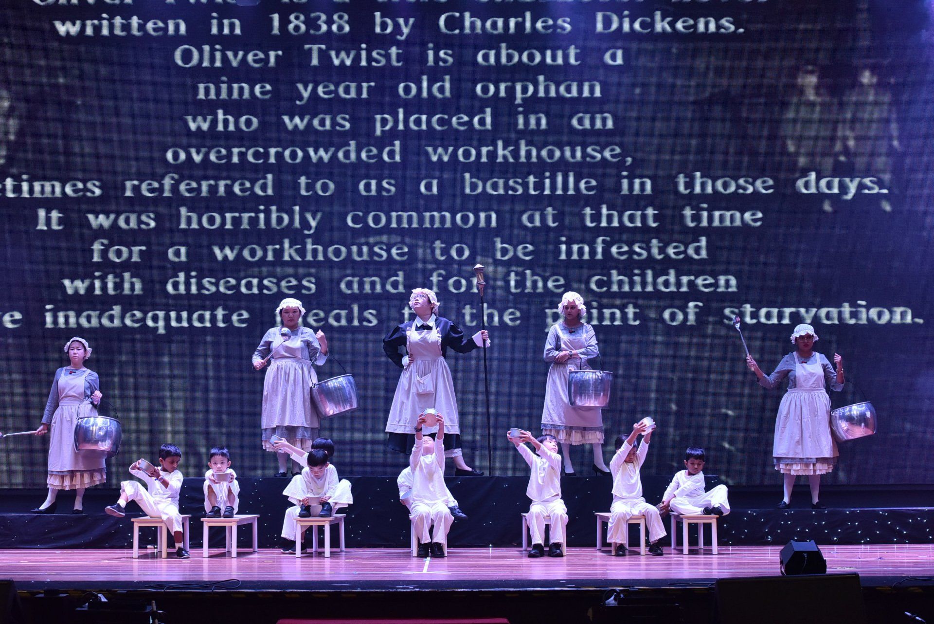 Children in costumes on stage, reenacting a scene from Oliver Twist, holding buckets. Projected text about the story.