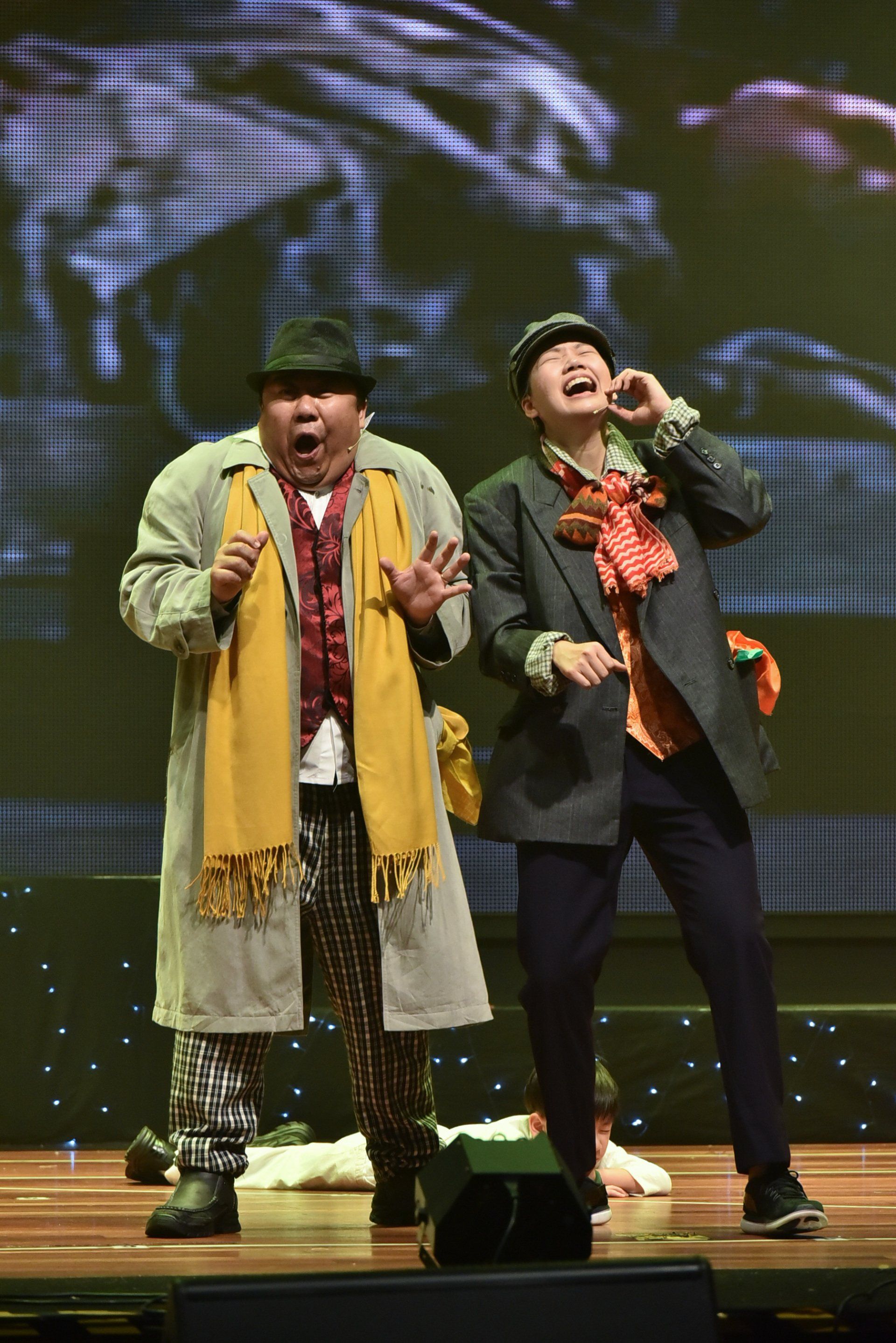 Two actors on stage, one in a trench coat and scarf, the other in a suit, both with expressive faces.
