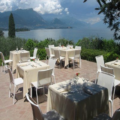 Tavoli da pranzo all'aperto in riva al lago con sedie bianche, con vista sulle montagne e un cielo nuvoloso.