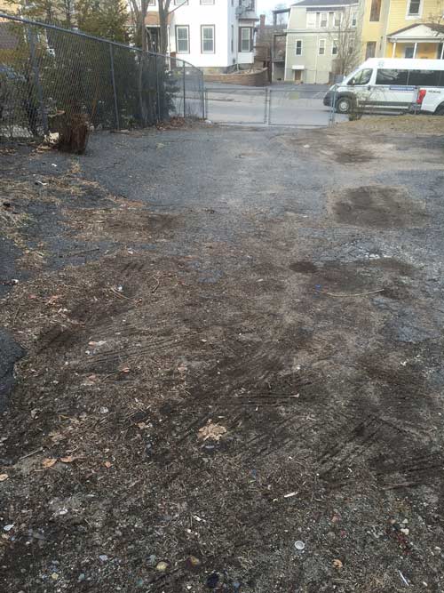 Gravel parking area; chain-link fence on the left; buildings in background. A van is parked to the right.