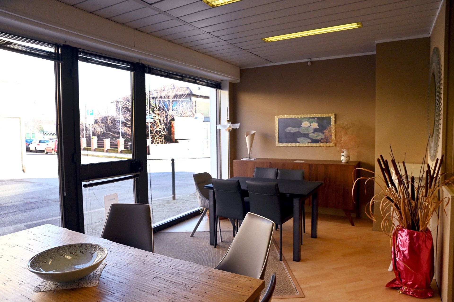 Area vuota del bar con tavoli, sedie e un vaso rosso accanto alle grandi vetrine.