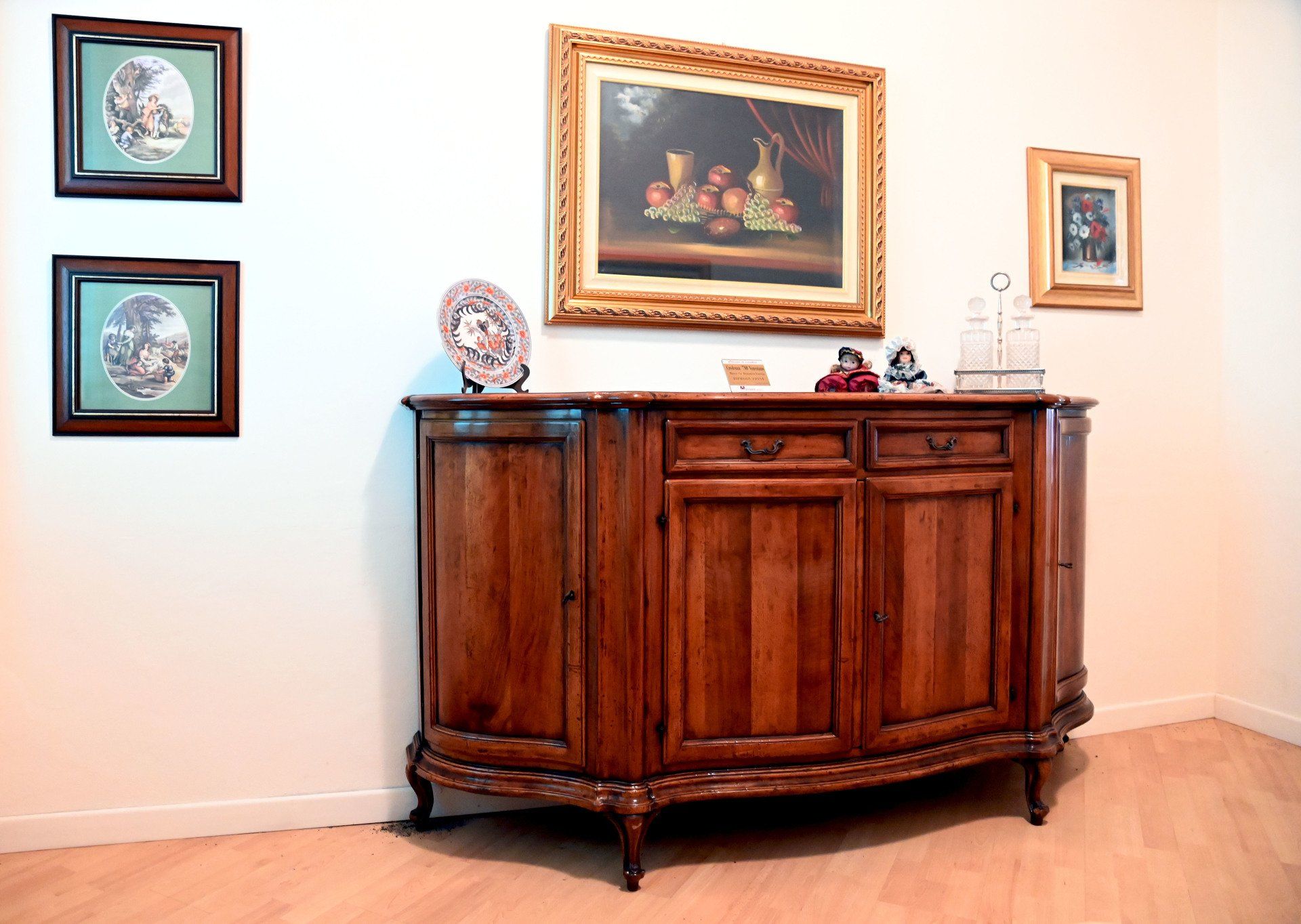 Una credenza in legno sotto un quadro incorniciato in una stanza luminosa e vuota con pavimento in legno chiaro.