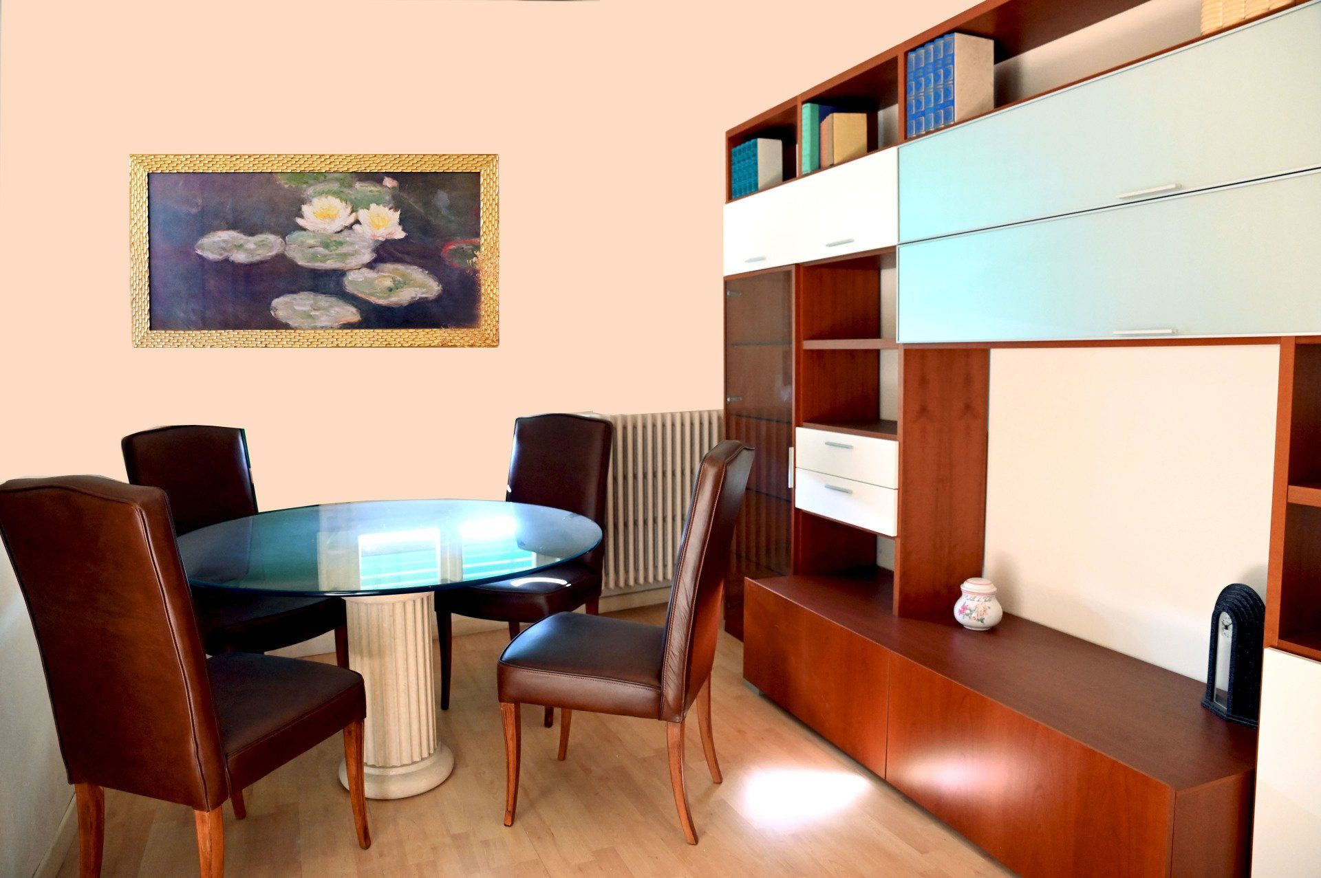 Sala da pranzo moderna con tavolo rotondo in vetro, sedie marroni, quadri alle pareti e scaffalatura a muro.