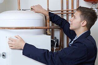 Working On Central Heating Boiler - Plumbing Repair in Yorkville, IL A young man in a navy jacket is working on a white water heater, using a wrench. Copper pipes are visible in the background.