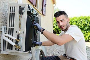 Air Condition Repair- Plumbing Repair in Yorkville, IL Man in gloves repairing outdoor air conditioning unit. He's looking at the camera, in a residential setting.