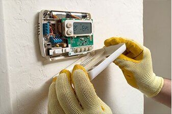Thermostat Repair - Plumbing Repair in Yorkville, IL Person wearing yellow gloves removing a thermostat cover on a white wall. The thermostat's electronic components are exposed.