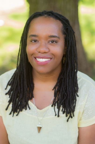 A woman with dreadlocks is wearing a yellow shirt and smiling.