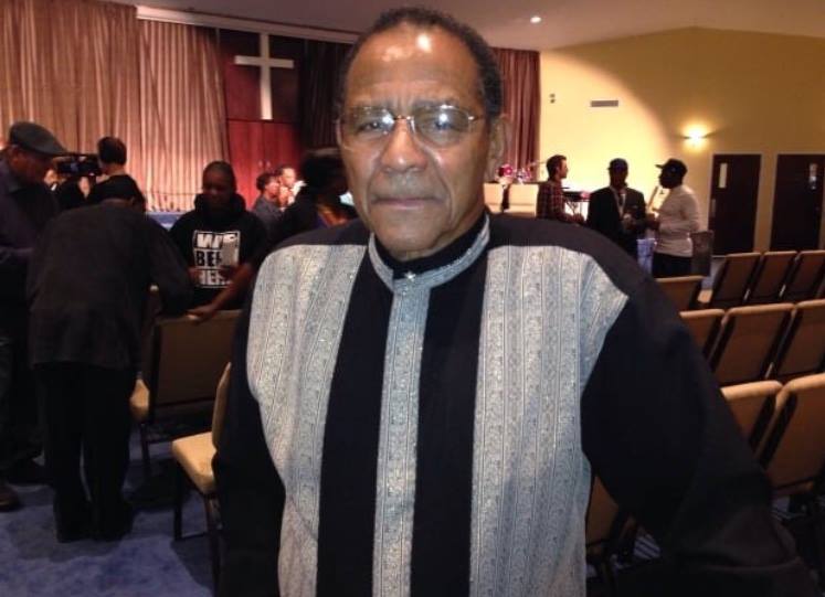 A man wearing glasses stands in a church with a cross in the background