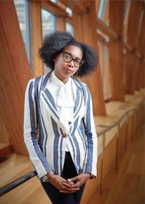 A woman wearing glasses and a striped jacket is standing in front of a window.