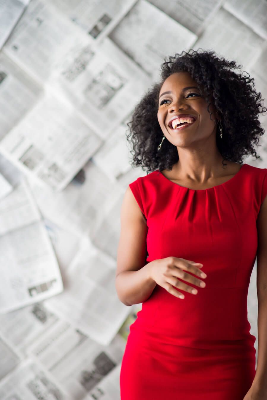 A woman in a red dress is standing in front of a wall of newspapers.