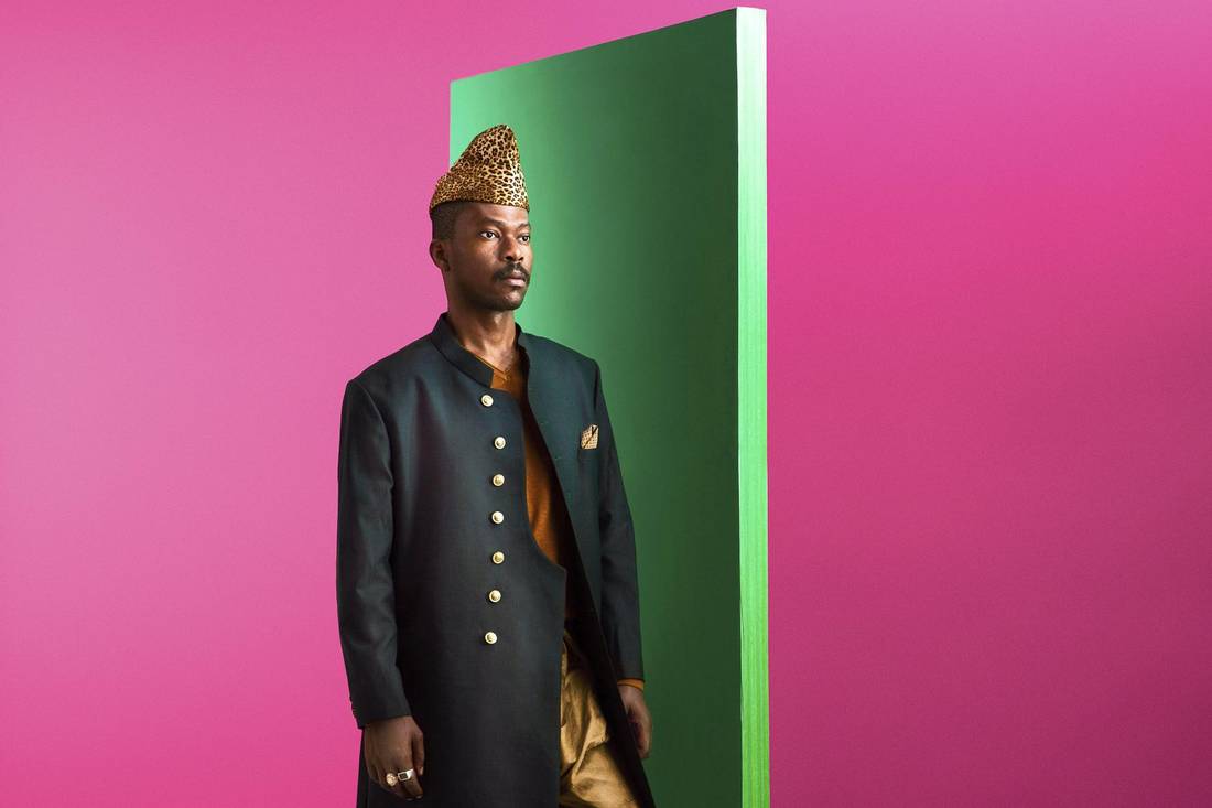 A man in a suit and hat is standing in front of a green wall.