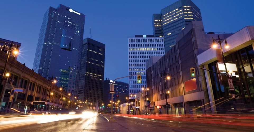 A city street at night with a lot of buildings and cars driving down it.