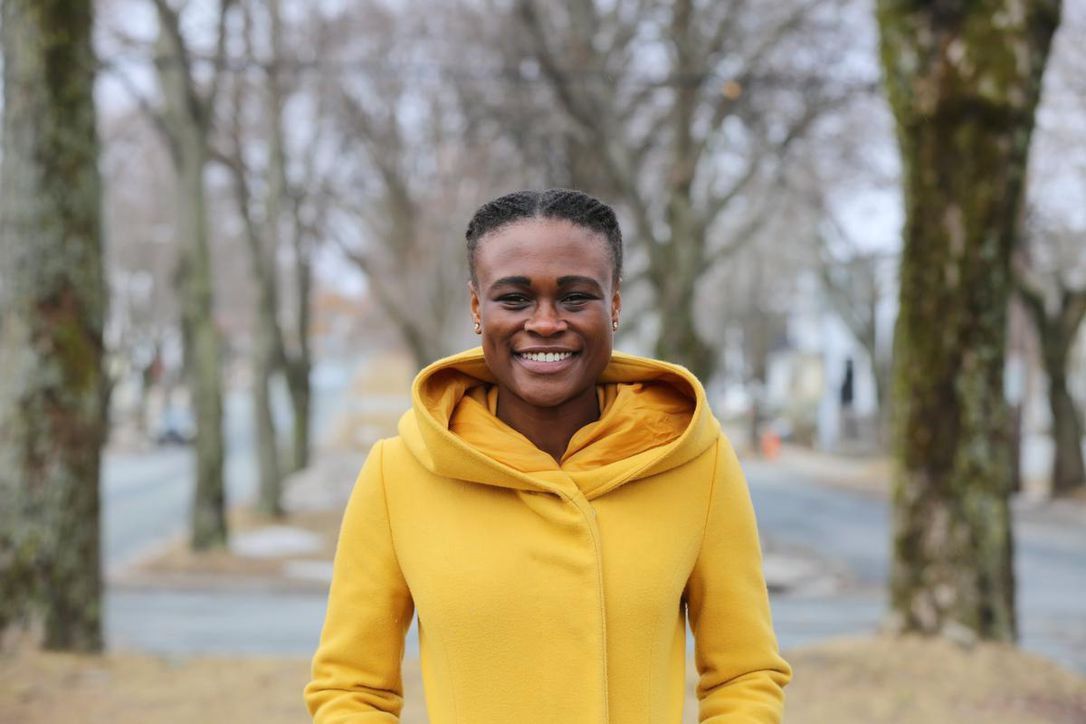 A woman in a yellow coat is standing in front of trees.