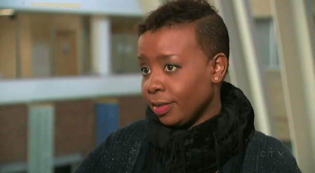 A woman with a shaved head is wearing a black sweater and scarf