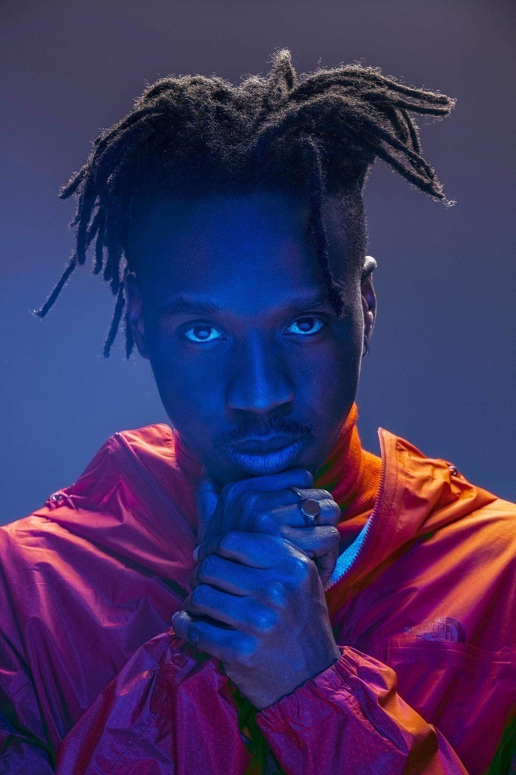 A man with dreadlocks is wearing a red jacket and holding his hands together.