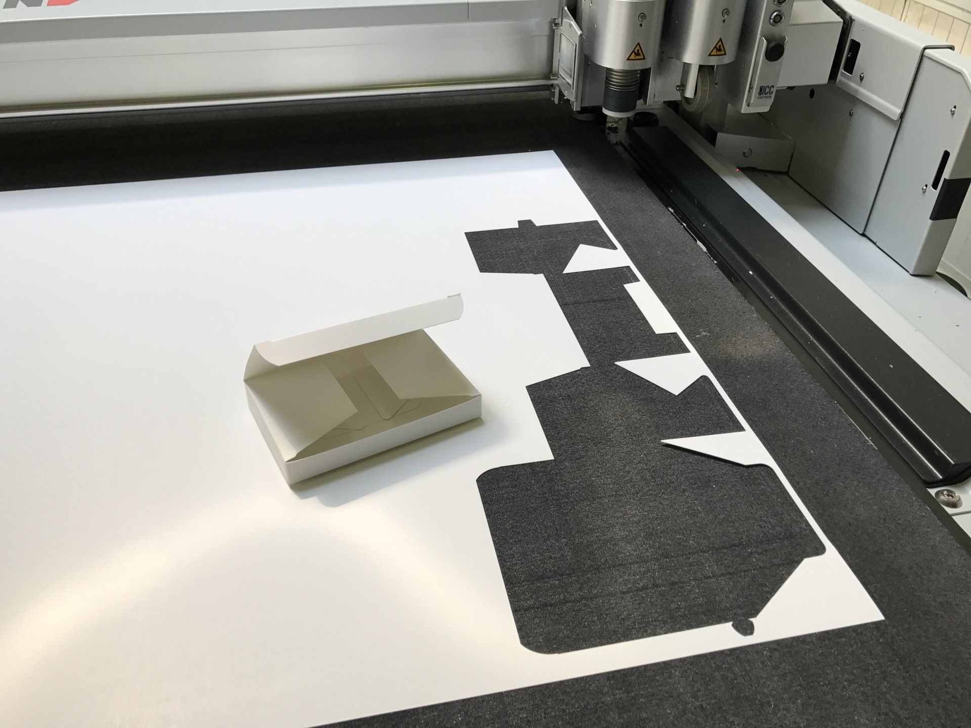 Een witte kartonnen doos naast een snijmachine op een wit vel papier. Zwarte vormen zijn uit het vel papier gesneden.