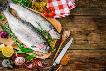 Dos peces están sentados sobre una tabla de cortar de madera rodeados de verduras y especias.