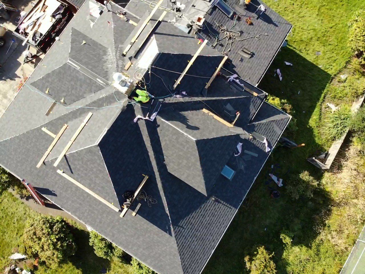 Roof under construction; workers on dark shingled roof with wooden supports. Sunlight, green grass visible.