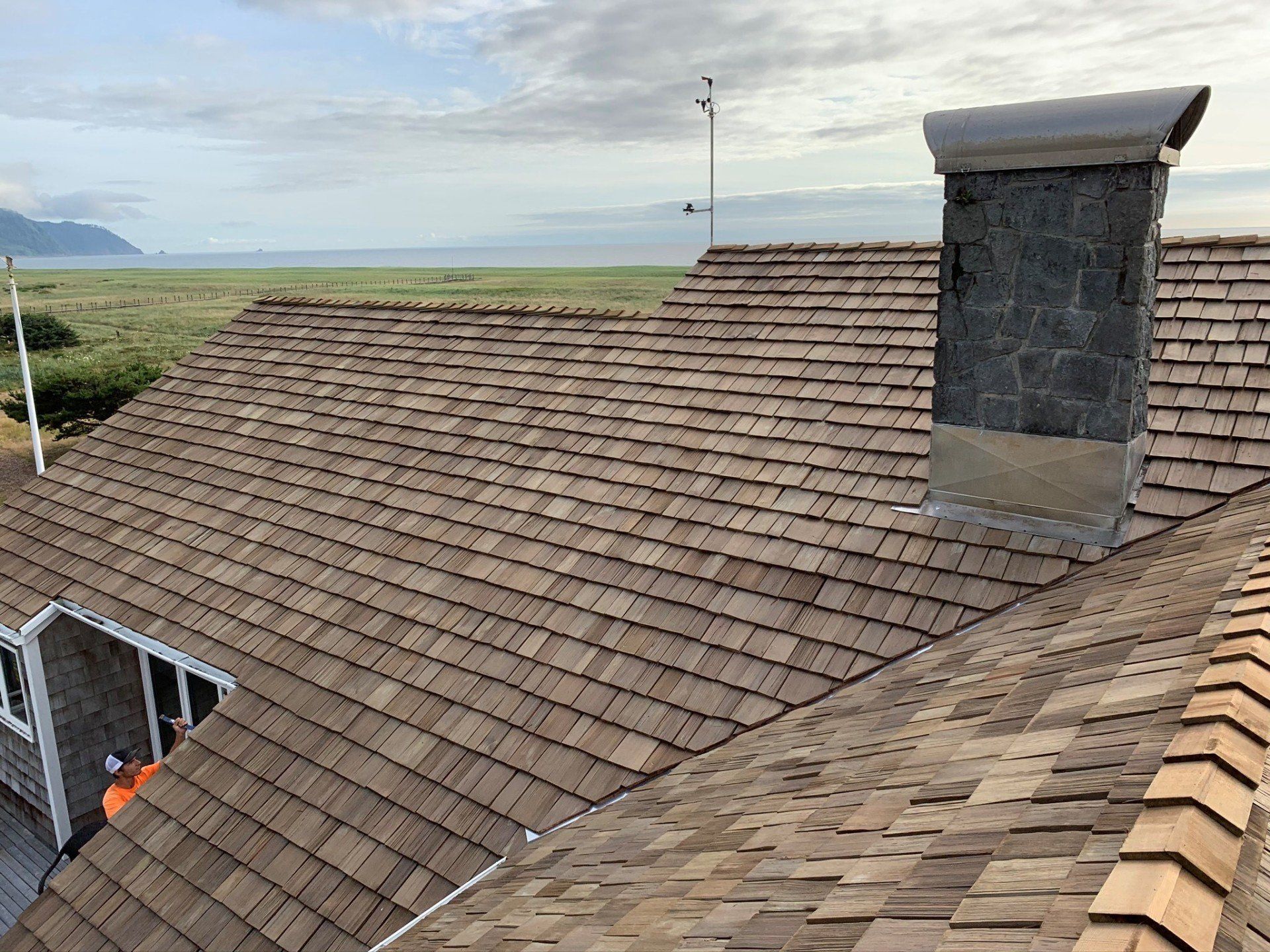 Brown shingle roof with chimney against a backdrop of green field and cloudy sky.