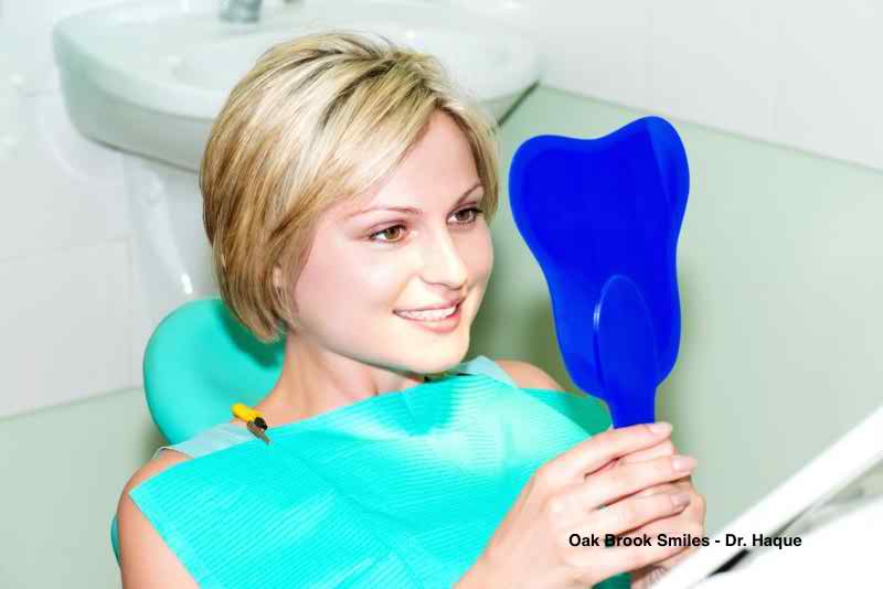 Woman in a dental chair looking at a blue tooth-shaped mirror, smiling. Green drape, white sink visible.