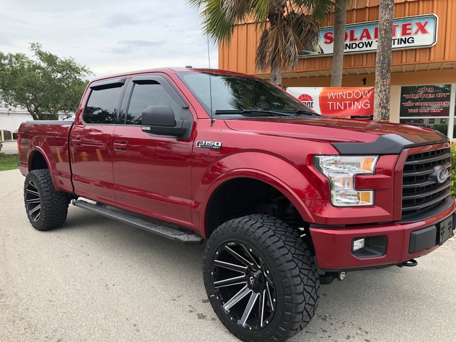 Red Ford truck with black wheels parked in front of a storefront with palm trees in the background.