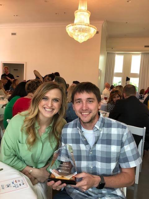 Smiling couple at a banquet, man holds an award. Woman wears green, man in plaid, chandelier overhead.