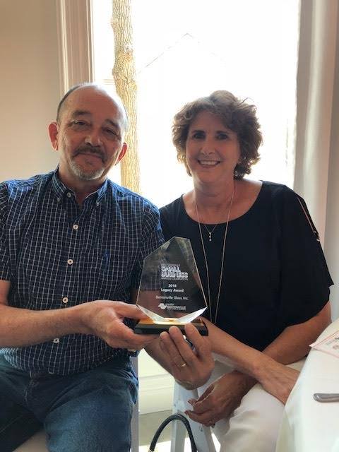 Man and woman holding an award, smiling. He wears a blue shirt, she wears black. Bright setting.