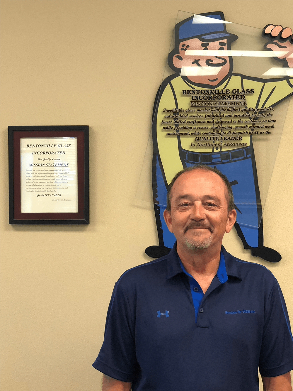 Man smiling in a blue shirt stands in front of a wall with a framed document and a cartoon character graphic.