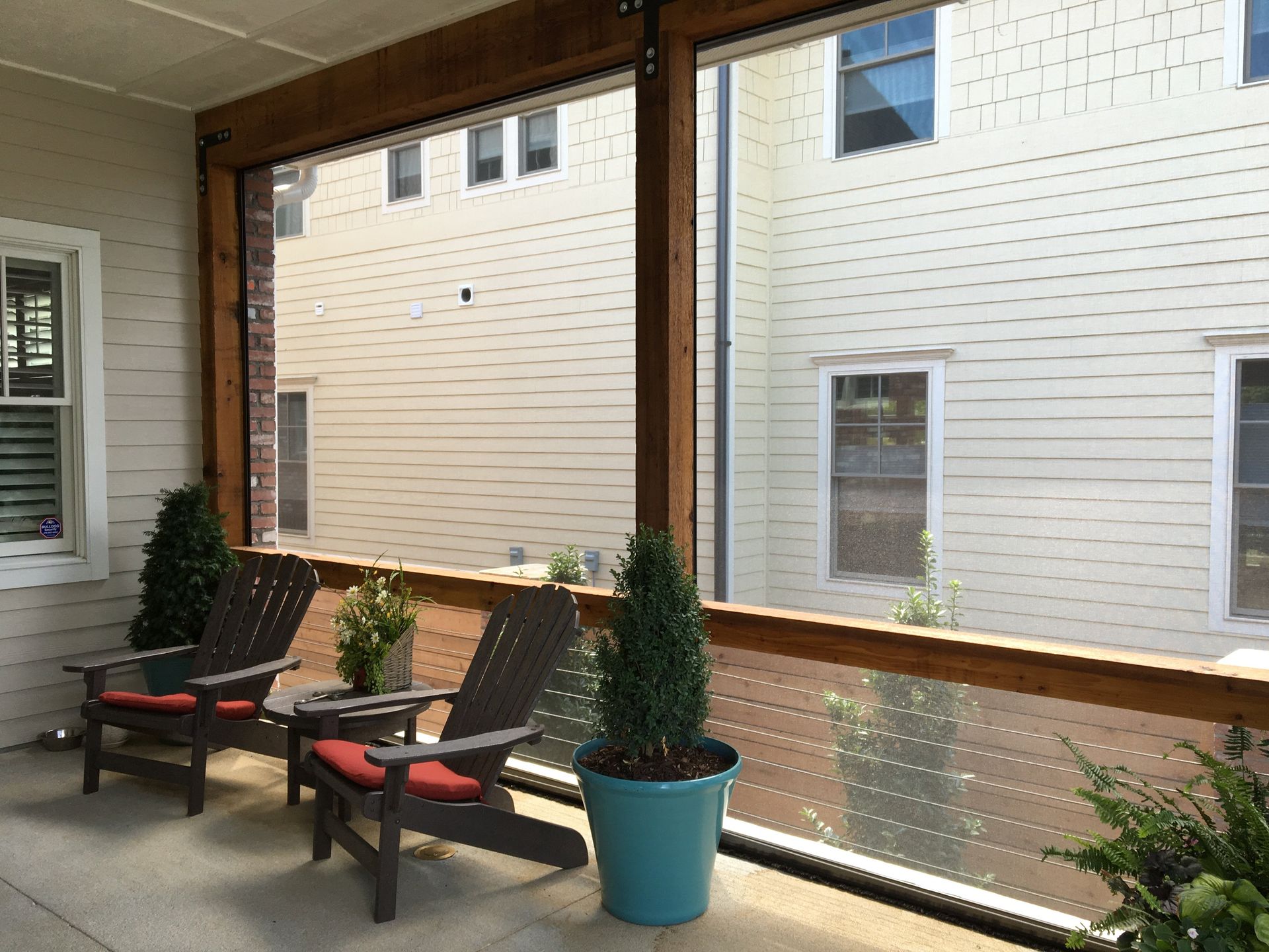 Covered porch with two wooden chairs, potted plants, and clear screens overlooking a building.