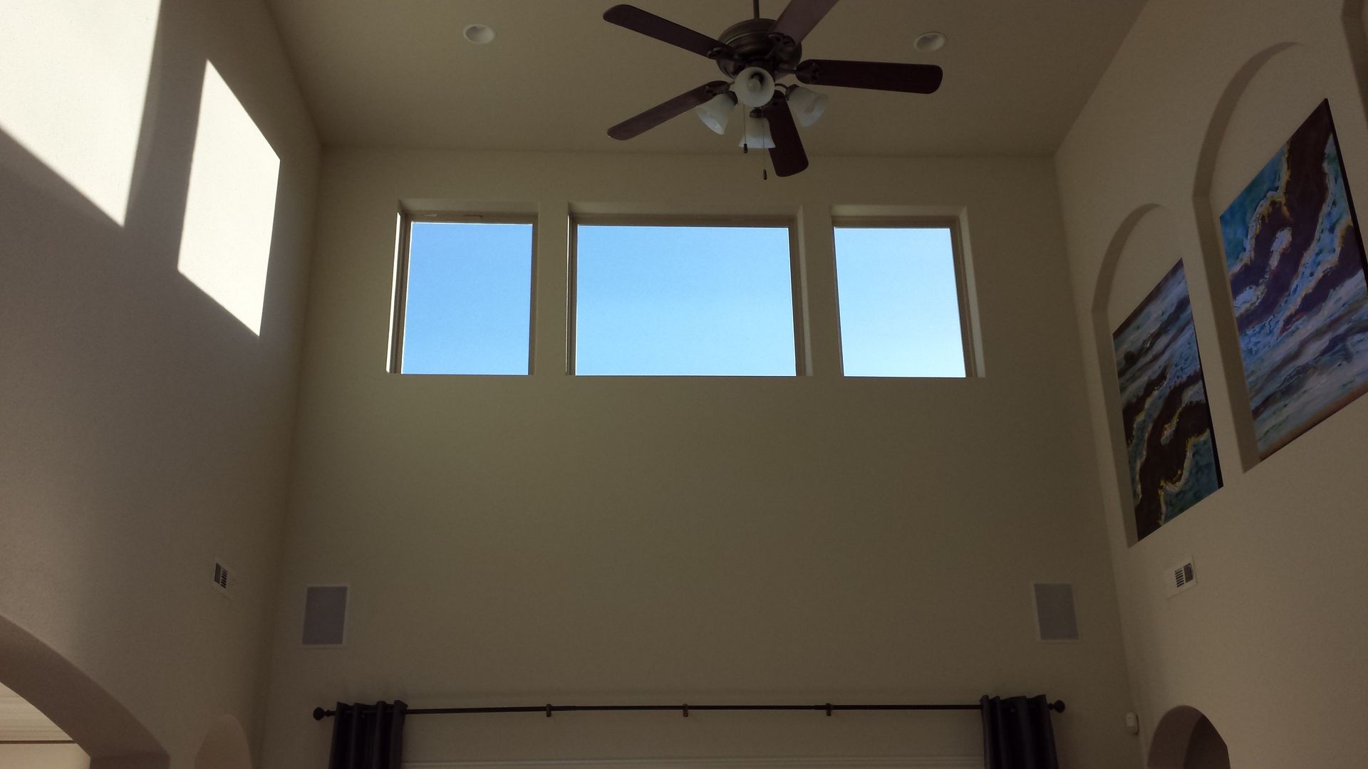 High-ceilinged room with three windows, a ceiling fan, and art on the wall, lit by sunlight.