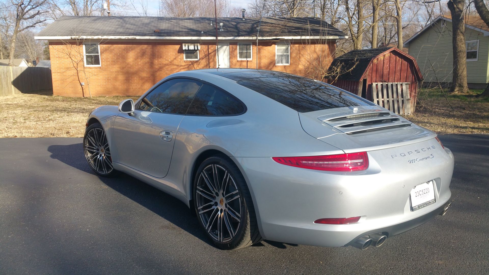 Silver Porsche 911 parked on a driveway in front of a modest, single-story house and a shed.