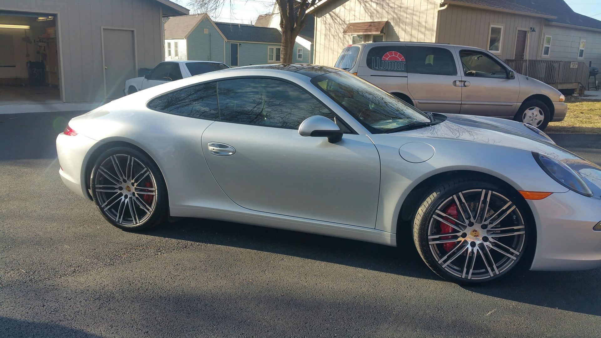Silver Porsche parked on a driveway, red brake calipers visible. A garage and houses in the background.