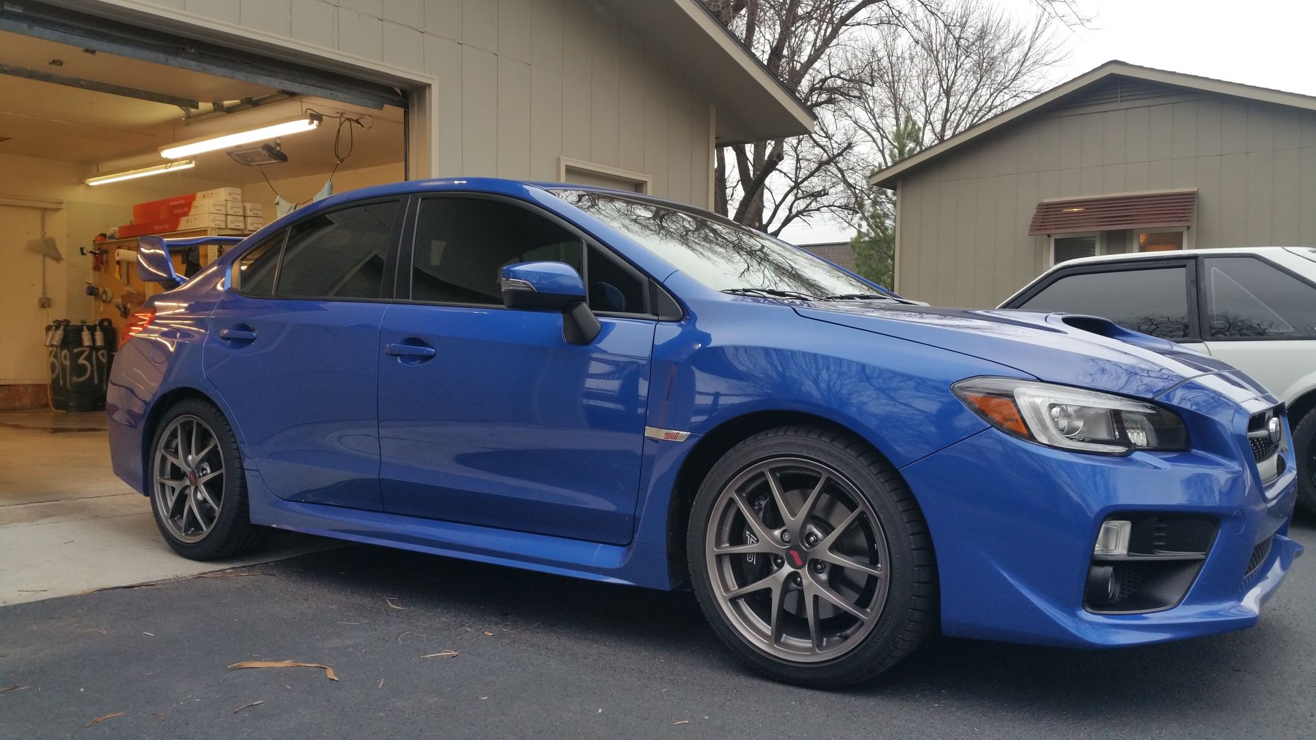Blue Subaru WRX sedan parked in front of a garage, with a white car visible in the background.