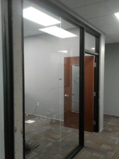 Glass-walled office space with brown door. Gray walls, tiled floor, and overhead lights.